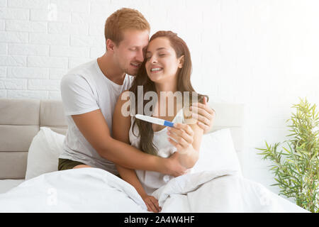 Giovane uomo è felice costeggiata bella donna sorridente quando ella lo mostra al test di gravidanza mentre entrambi sono seduti a letto Foto Stock