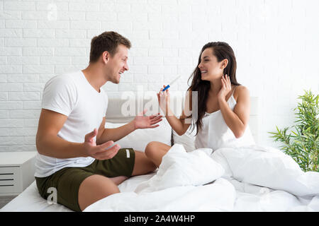 Giovane uomo è felice quando bella donna sorridente lo mostra al test di gravidanza mentre entrambi sono seduti a letto Foto Stock