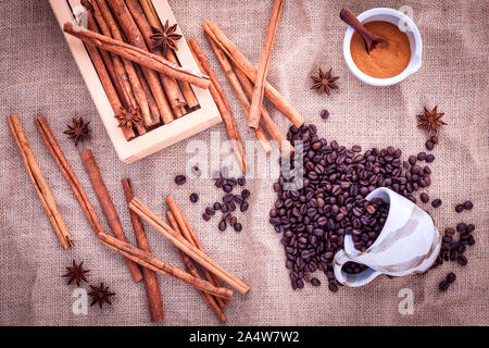 La tazza di chicchi di caffè sul panno del sacco con bastoncini di cannella ,cannella in polvere nel recipiente e anice stellato. Foto Stock