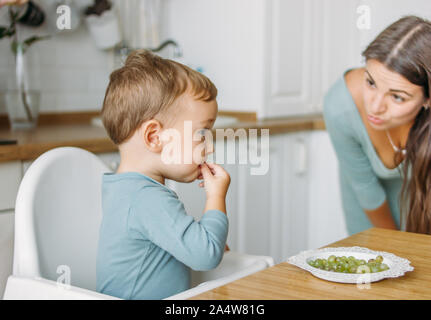 Affascinante piccolo concentrato baby boy mangiare cibo prima uva verde presso la luminosa cucina a casa Foto Stock