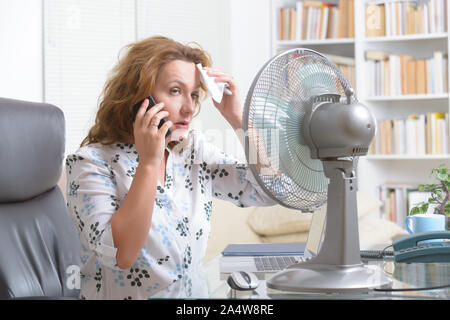 La donna soffre di calore mentre si lavora in ufficio e si tenta di raffreddarsi dalla ventola Foto Stock