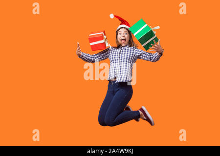 Ritratto di estremamente felice brunette donna in santa hat e camicia a scacchi salti di gioia e di volare con avvolto xmas confezioni regalo, celebra vendite. i Foto Stock