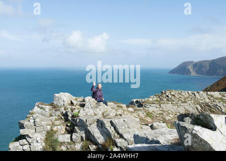 Donna walker sventolare alla telecamera seduta alta su una cengia rocciosa al di sopra del mare nella valle di rocce, Lynton, Devon. Foto Stock