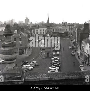 1965, storica, vista dall'alto della piazza del mercato con auto parcheggiate, Alyesbury, Bucks, Inghilterra, Regno Unito, mostra l'architettura, gli edifici pubblici e i negozi dell'epoca, tra cui la sartoria di Burton. Al centro della piazza, la torre dell'orologio con la guglia. Costruito nel 1876, è stato progettato dall'architetto D. Brandon, che ha anche progettato il Corn Exchange. Foto Stock