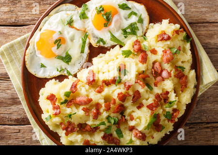 Gustosi piatti caldi di purè di patate con pancetta e prezzemolo e servite con uova fritte close-up su una piastra sul tavolo. Parte superiore orizzontale vista da sopra Foto Stock