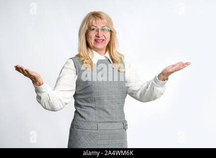 Donna matura mostra le sue mani in direzioni differenti, in piedi su sfondo grigio. Successo donna senior. Insegnante. Foto Stock