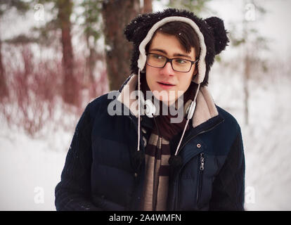 Carino affascinante ragazzo sorpreso in una giacca blu, cappello con orecchie e cuffie bianche si erge nel mezzo di un inverno di foresta di pini e sembra sorpreso. Foto Stock