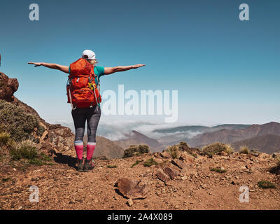 Gioiosa ragazza turistica permanente sulla cima della montagna in Alto Atlante del Marocco e guardando in giù per la valle con le nuvole Foto Stock