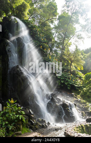 Cascata con luminosi raggi del sole lo scoppio attraverso la nebbia creata dalla caduta di acqua al Parque Natural da Ribeira Caldeirões dos, São Miguel. Foto Stock