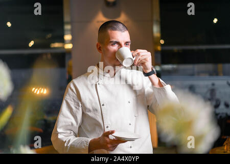 Bel giovane cuoco capo di bere il caffè del mattino dal bianco cup nel lusso moderno ristorante interno. Foto Stock