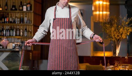 Bel giovane cuoco capo grembiule di legatura nel moderno ristorante interno. Close up Foto Stock