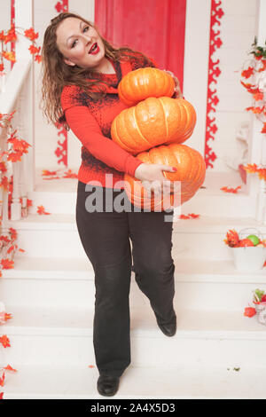 Chubby giovane donna caucasica in arancione maglione holding zucche permanente sulla veranda della sua casa bianca Foto Stock