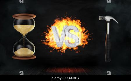 3D rendering di un calcestruzzo flaming lettere VS stand tra una clessidra completa e un martello industriale. Foto Stock