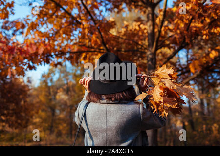 La stagione autunnale. Giovane donna camminare nel parco di caduta ammirando la natura e la holding bouquet di foglie. Vista posteriore Foto Stock
