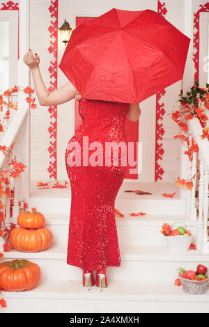 Sinuosa donna in una lunga e luminosa abito da sera si erge sulla veranda della sua casa bianca con la schiena e tiene aperto un ombrello rosso. Foto Stock