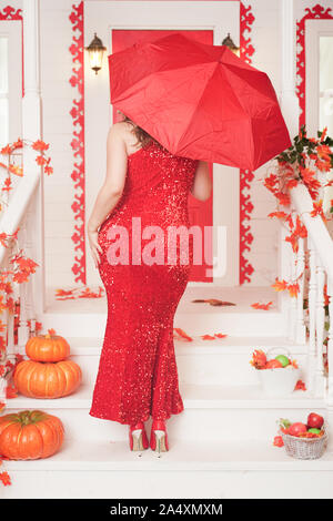 Sinuosa donna in una lunga e luminosa abito da sera si erge sulla veranda della sua casa bianca con la schiena e tiene aperto un ombrello rosso. Foto Stock