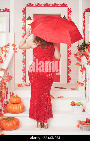 Sinuosa donna in una lunga e luminosa abito da sera si erge sulla veranda della sua casa bianca con la schiena e tiene aperto un ombrello rosso. Foto Stock