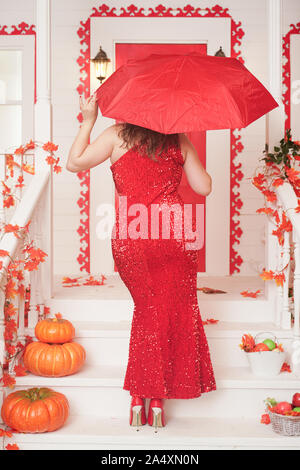 Sinuosa donna in una lunga e luminosa abito da sera si erge sulla veranda della sua casa bianca con la schiena e tiene aperto un ombrello rosso. Foto Stock
