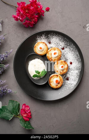 Frittelle con panna acida in una piastra nera decorata con zucchero a velo su sfondo grigio Foto Stock