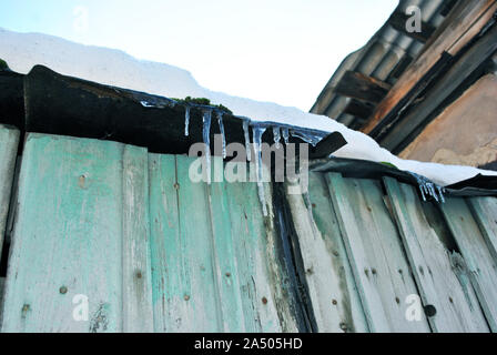 Legno verde vecchio muro con scuri malandato tetto con ghiaccioli e il bianco della neve sulla parte superiore, vista dal suolo su blu cielo chiaro sfondo Inverno nel paese Foto Stock