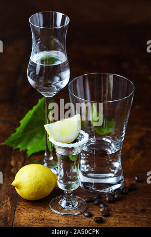 Elevato angolo di close-up di grappa bianca scatti in tre diversi vetri trasparenti, decorata con limone, i chicchi di caffè e uva verde foglie sul buio la Foto Stock