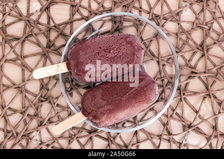 Vista superiore crudo fresco popsicles su bastoni rivestiti con glassa di cioccolato laici in vetro ciotola trasparente sulla tovaglia in vimini. Deliziosi piatti vegan in casa Foto Stock