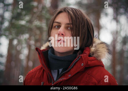 Fiducioso un bel ragazzo in giacca rossa vale la pena in mezzo a pini bosco invernale e sembra freddo mournfully heartless. Foto Stock