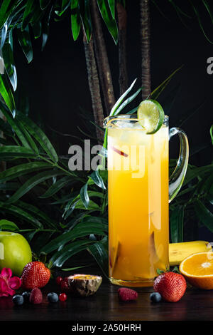 Ananas mango cocktail in una brocca su sfondo scuro con frutti: ananas, mango, confettura di fragole, purea di mango, succo di ananas, pesca, mela, Sprite Foto Stock