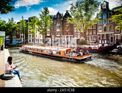 Giovane donna seduta sulla riva del canale guardando una piacevole gita in barca attraverso i canali di Amsterdam, Olanda, Europa. Foto Stock