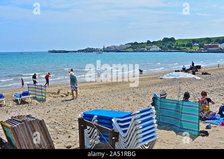 Spiaggia tradizionale scena con famiglie godendo del sole & paddling in mare. Swanage pier in background. Gruppo familiare sotto ombrellone & con frangivento. Foto Stock