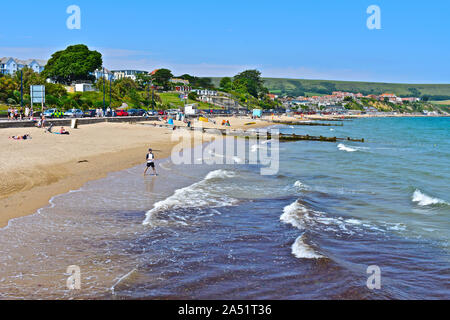 Onde arrotolate delicatamente in spiaggia di sabbia a Swanage Bay. Le persone che si godono il sole estivo. Pennelli in legno azienda sabbia in luogo.case sulla collina alle spalle. Foto Stock