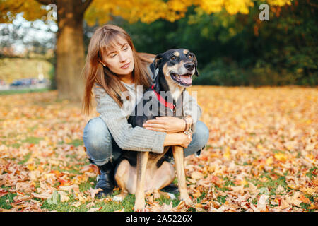 Ritratto di bella felice caucasico giovane donna seduta su un terreno tra autunno cadono le foglie giallo e abbracciando il suo cane. Migliori amici divertendosi o Foto Stock