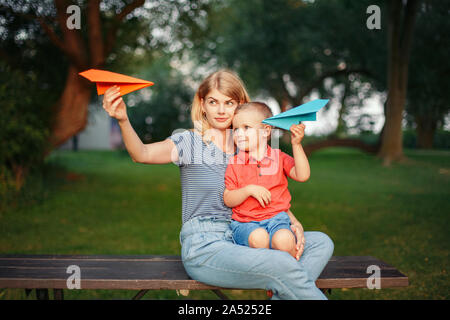 Giovane madre caucasica e ragazzo toddler figlio seduti insieme all'esterno sul tramonto estate giocando con carta colorata aerei. La famiglia felice infanzia Foto Stock