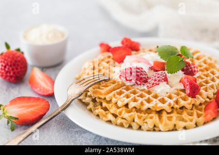 Fresche e dorate di cialde con un trito di fragole e panna montata sulla parte superiore. Piastra bianca e sfondo grigio e un pezzo di tessuto bianco accanto alla piastra. Foto Stock