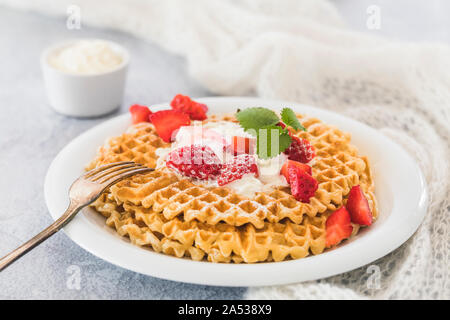 Una piastra con una pila di fresco cialde dorata con un trito di fragole e panna montata sulla parte superiore. Piastra bianca e sfondo grigio e un pezzo di bianco fa Foto Stock