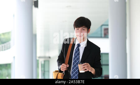 Bello sorridente giovane imprenditore tuta da indossare all'aperto a piedi tenendo tazza di caffè durante la pausa caffè. Foto Stock