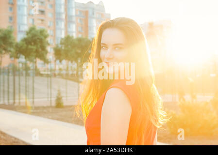 Luce posteriore ritratto di un felice sorridente giovane donna, business lady in tuta rossa al tramonto in un parco con una calda luce gialla e background urbano. Selezione Foto Stock