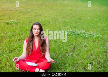 Giovane imprenditrice in tuta rossa seduta nella posizione del loto su erba verde nel parco e tenendo il suo smart phone. Meditazione per LFE. Calma, rilassata lifest Foto Stock