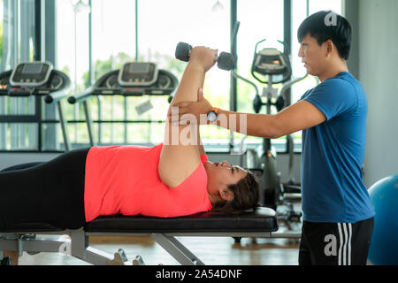 Due Asian trainer uomo e donna sovrappeso che esercitano con il manubrio insieme in una palestra moderna, felice e sorridere durante gli allenamenti. Grasso donne prendersi cura di hea Foto Stock