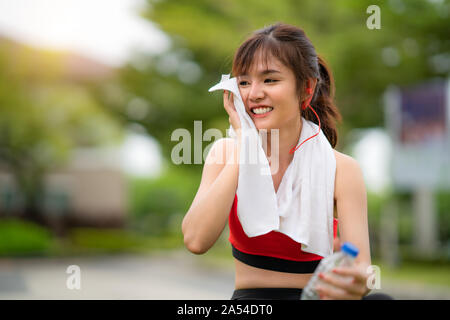 Asian bella donna seduta per rilassarsi dopo allenamento da soli e trattenere acqua in bottiglia in parco pubblico nel villaggio, felice e sorriso in mattinata durante sunl Foto Stock