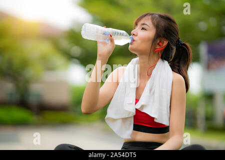 Asian bella donna seduta per rilassarsi dopo allenamento da soli e acqua potabile dalla bottiglia in parco pubblico nel villaggio, felice e sorriso in mattinata assortiti Foto Stock