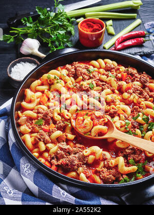 Gulasch di gomito pasta, carni bovine, il sedano e i pomodori in un nero piatto di ceramica su un tavolo di legno con carta asciugatutto, vista da sopra Foto Stock