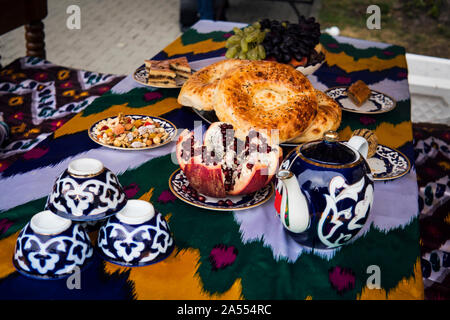 Tradizionale pane uzbeko sul tavolo con tazze e bollitore. La cucina di Asia centrale. frutta uva e melograno e dolci in piastre Foto Stock