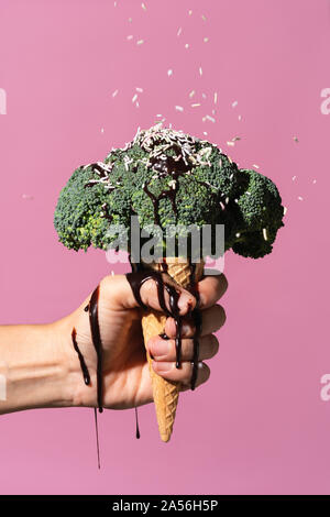 Studio colpo di mano d'uomo tenendo cono gelato con broccoli in cima, gocciolamento in salsa di cioccolato e spruzza, contro sfondo rosa Foto Stock