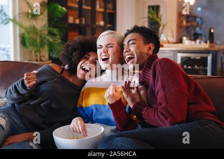 Giovani uomini e giovani donne amici di ridere mentre si guarda la TV sul divano di casa con una ciotola di pop-corn Foto Stock