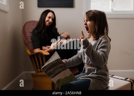 Ragazza con libro facce di trazione, mentre madre culle baby fratello in salotto poltrona Foto Stock