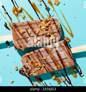 Due fatti in casa fudge lollies ghiacciata con spruzzata e spruzzata dadi, caramello e salsa al cioccolato su sfondo blu, vista aerea Foto Stock