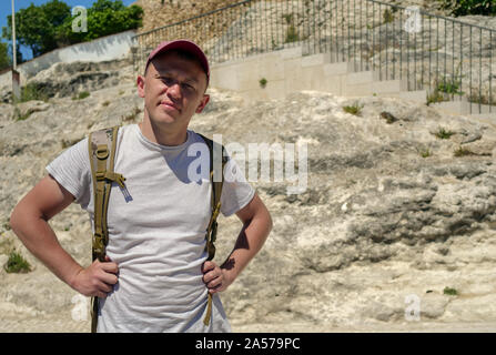 Ritratto di un giovane maschio turistico con uno zaino sulla strada nei pressi di una roccia di pietra con una foresta, guardando direttamente verso la telecamera Foto Stock