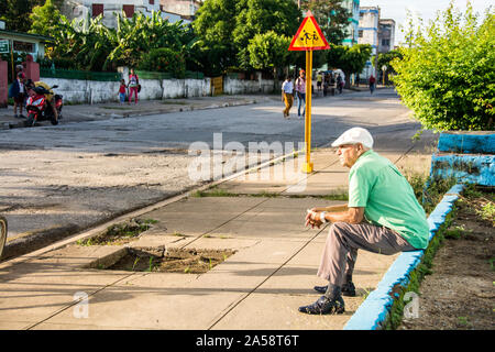 Un vecchio cubano seduto sul fianco di un marciapiede; Camaguey, Cuba Foto Stock