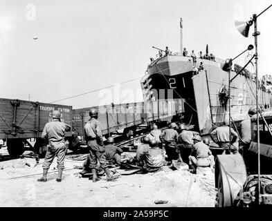 [Didascalia originale: ]I GUARDACOSTE LINKS britannica e francese ferrovie: fuori del massiccio di ganasce di un U.S. Coast Guard-presidiati (LST) (sbarco navi, cisterne) rotoli di una stringa di vagoni ferroviari, caricato con vitale bellica in Inghilterra e consegnati in Francia. Il Coast Guard-presidiati nave Traghetti materiale rotabile da un porto britannico di una spiaggia francese del fungo, formando un collegamento mobile tra le ferrovie dei due paesi. Ore di inestimabile valore, lo scarico e il ricarico, vengono salvati eseguendo il pranzo di auto in pista attrezzata tank deck del LST e il trasporto attraverso il canale della Manica a rotolare giù per un Foto Stock